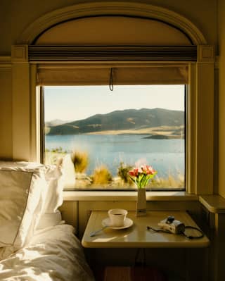 A coffee cup, a camera and a bud vase rest on a window table with a lake view in morning sun, next to an unmade bed.