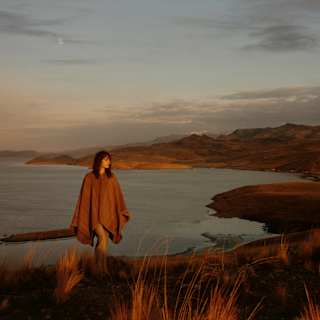 A woman in a brown wool poncho strides up an incline with a backdrop of hill chains and calm lake waters, seen at sunset.