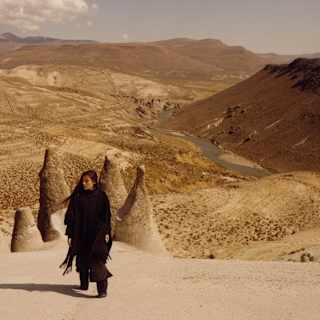 A woman with long dark hair and black clothes stands on a hill overlooking the Puruña stone forest and a valley river beyond.