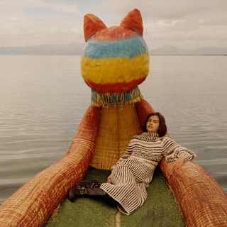 A dark-haired woman in a striped dress lies back on a Lake Titicaca boat made of reeds with a large cat-like figurehead.