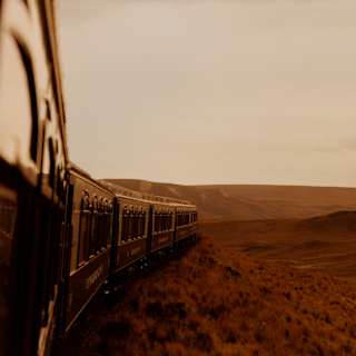 External view from the fifth carriage window capturing the train front forging through grasslands tinted deep-red by sunset.
