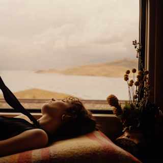 A woman reclines on bed by the window, holding a sash from her top as soft sun plays on her face and lake contours behind.