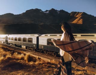 A Peruvian woman stands in the grass alongside the train track, watching the Andean Explorer pass, with the wind in her hair.