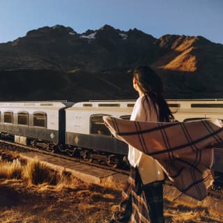 A Peruvian woman stands in the grass alongside the train track, watching the Andean Explorer pass, with the wind in her hair.
