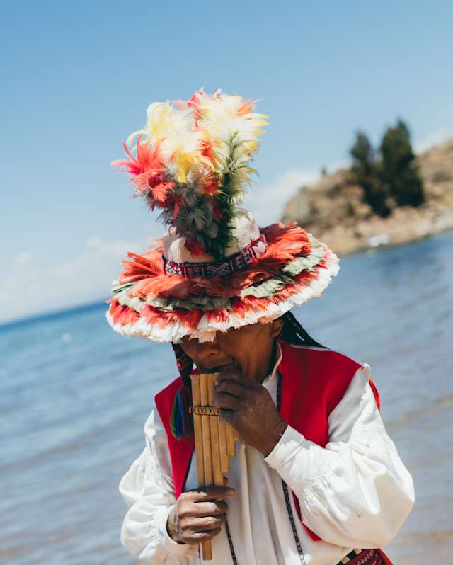 A man in local costume and a wide-brimmed hat erupting with red, cream and green feathers, plans panpipes by Lake Titicaca.