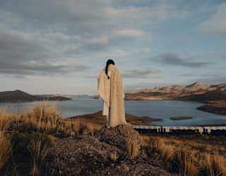 A woman wearing a white woollen shawl watches the train from a high rock as it passes Lake Saracocha below, seen from behind.