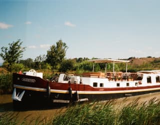 The Alouette sails the smooth brown waters of the canal in pleasant countryside beneath blue sky, seen over a grassy bank.