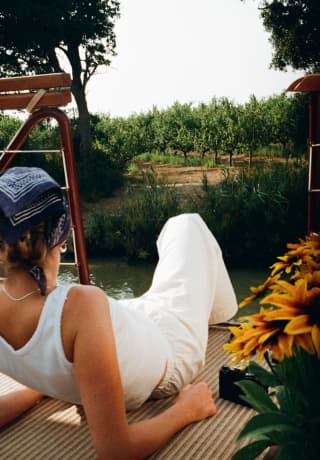 A woman in a white top and trousers with a blue headscarf lies back on the deck, gazing at the countryside, seen from behind.