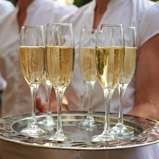A waitress holds a silver tray with six flutes of sparkling champagne, as other wait staff stand by, ready to serve.