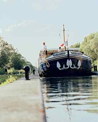 Water-level view of the Amaryllis on a narrow section of the Canal du Centre, about to be moored by a man holding a rope.