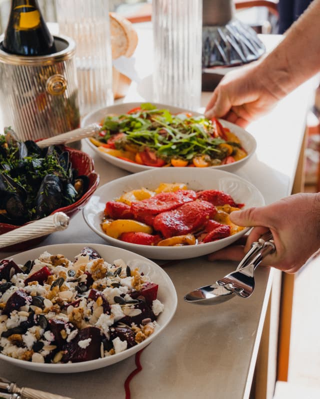A guest reaches for bowls of roasted capsicums and bright tomato salad at a buffet table with dishes, bread and champagne.