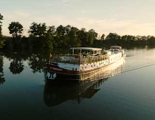 Evening sun brushes the side of the Coquelicot barge as she cruises along a calm waterway that reflects the tree-lined bank.