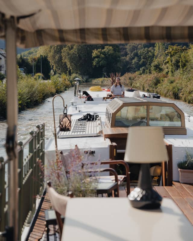 A crew member steers the barge, seen in a view from the upper deck along the wood and glass top of the Coquelicot boat.