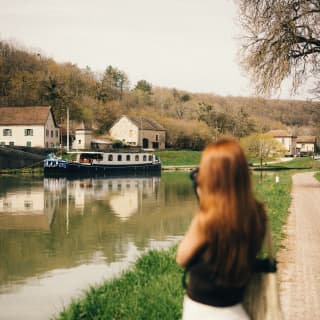A woman stands on the bank of the canal, taking a photograph of the Fleur de Lys as it cruises by a cluster of white houses.