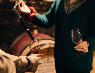 By a row of oak wine barrels in the cellar of a Burgundy vineyard, an expert pours red wine into a glass for a guest to try.