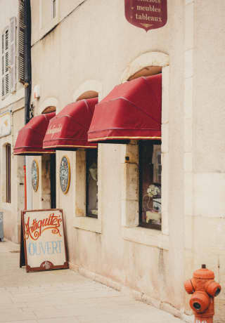 A hand-painted sign on the pavement outside a quaint antiques shop in Burgundy, with white walls and red window awnings.