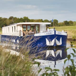The Lilas cruising with two passengers on deck enjoying the sunshine and tranquillity, pictured through grasses on the bank.