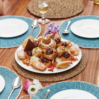 A piled plate of sugar-dusted Madeleines and choux buns with creme and chocolate rests on a laid table with blue place mats.