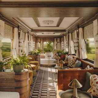 A luxurious vintage train carriage interior with large windows, cream curtains, patterned flooring, plush sofas, plants, and decorative cushions, creating an elegant and cosy atmosphere while the scenery blurs outside.