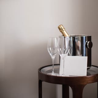 The foil neck of a Champagne bottle juts from a silver ice bucket on a small round table with two glasses and a welcome note.