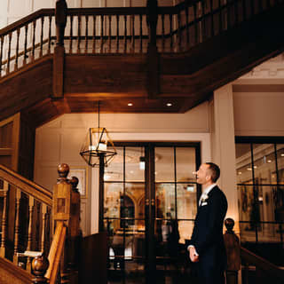 A dark-haired bride in a fitted gown holds a bouquet and smiles down at her groom from the top of a sweeping oak staircase