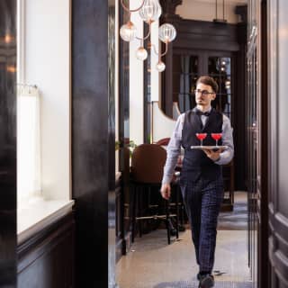 A waiter in dark waistcoat and bow tie walks below glass globe wall lights carrying a silver tray with two raspberry martinis