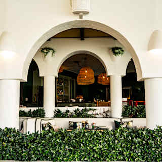 View through a white-rendered double colonnade to terrace seating and a restaurant behind, lit by wicker pendant lamps.