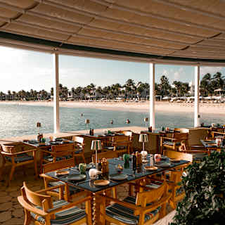 View across a dining terrace with wooden chairs and black marble-topped tables, laid for lunch, overlooking Maundays Bay.