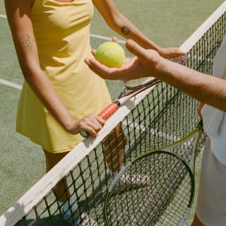 Close-up of a woman in a yellow dress and a man holding a tennis ball as they meet either side of the net with their rackets.