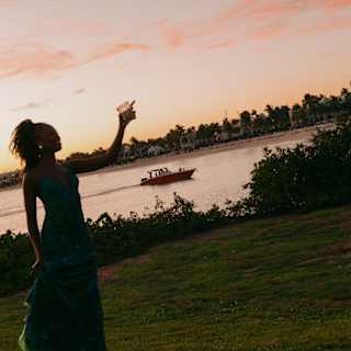 A woman holding up a drink is silhouetted against a pink sunset sky that tints the bay behind, where a small boat cruises by.