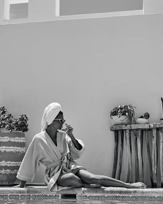 Black and white photo of a lady in a bathrobe posing on a poolside and sipping from a glass