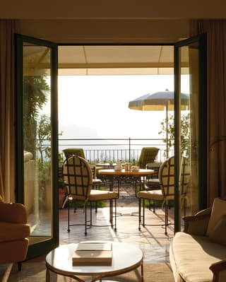 Hotel room lounge area with open patio doors and a sunlit balcony beyond
