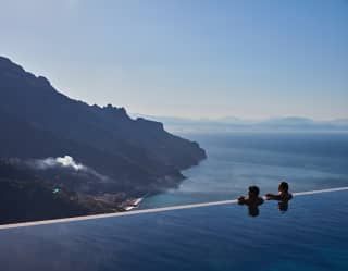 A man and a woman rest at the edge of an infinity pool, gazing at the view that plunges down steep cliffs below them