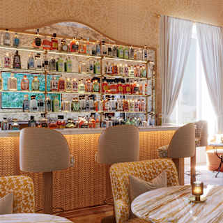 Ochre-veined marble tables with complementary tub chairs fill a bar lounge with a cane-fronted bar, stools and bottle shelves.
