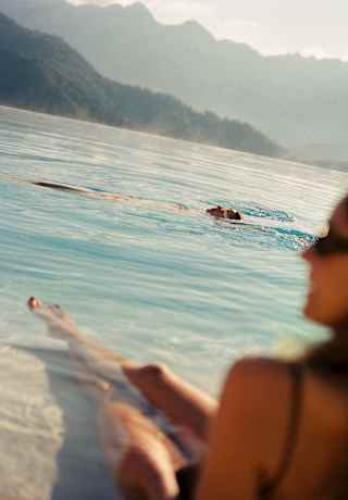 A man swims on his back in the pool, seen over the shoulder of a woman in soft-focus in an image that captures the hill views.
