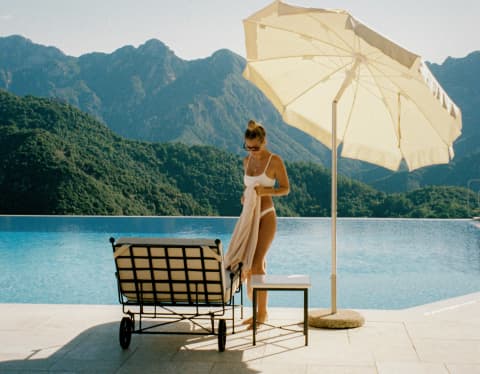 A woman in a white bikini stands with a towel next to her poolside lounger, beneath a parasol, with a mountainous backdrop.