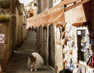A young girl dressed in a white Confirmation Dress bends to adjust her sock, shaded by an archway on a cobbled Ravello alley