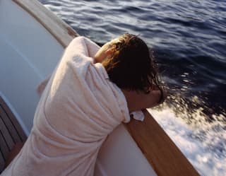 A woman, wrapped in a cream towel, rests her head on her folded arms on the side of a moving boat, watching the ocean ahead.