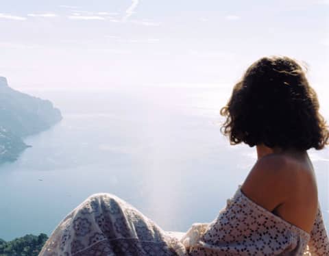 A woman with a dark bob, in a stunning strapless lace dress, turns her head to gaze at the sublime Amalfi coast views below.