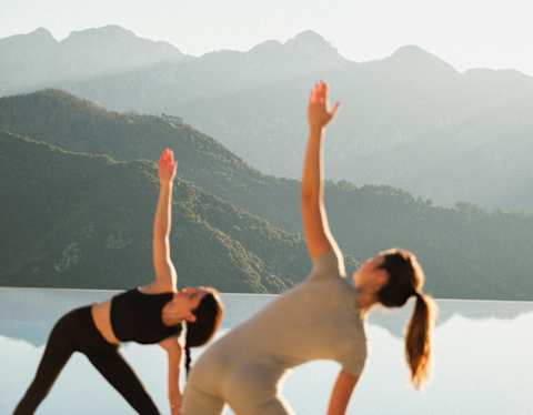 A yoga teacher in black and a guest in beige adopt the Extended Side Angle Pose, seen in soft-focus by the pool.