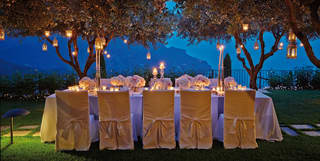 Lanterns hang from garden olive trees, illuminating a dining table set with flowers and candles for a wedding banquet