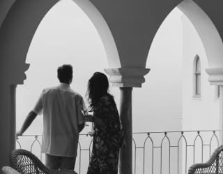 Black and white image of a couple looking out at the mist from their balcony, framed by wall arches and seen from behind.