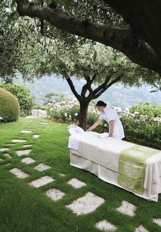 A masseuse sets up a massage couch in the shade of a tree in a flower garden with views of the Lattari Mountains>
