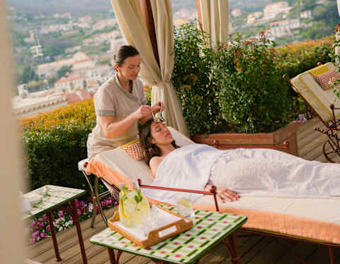 A spa therapist sits at the head of a treatment bed where she massages the forehead of a guest who lies beneath a sheet.