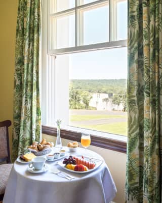 Breakfast including juice, pastries and fresh fruit on a white-clothed table by an open sash window overlooking a sunny rural view