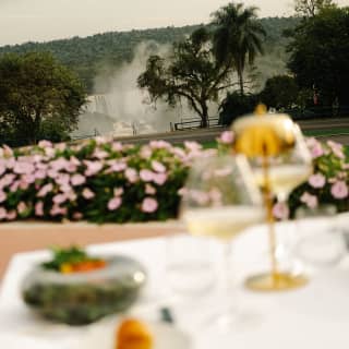 Cascading falls create clouds of spray in a garden view over a table with two glasses of white wine and food, in soft-focus.