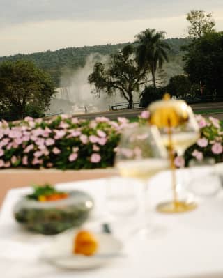 Cascading falls create clouds of spray in a garden view over a table with two glasses of white wine and food, in soft-focus.