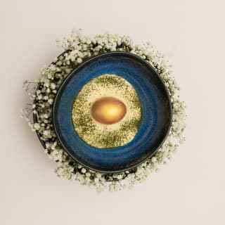 In a midnight blue bowl encircled by white flowers, a gold egg rests in a pool of seasoned tucupi sauce, seen from above.