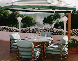 A large green-striped cantilever parasol shades a round table with four wine flutes and pale blue chairs overlooking gardens.