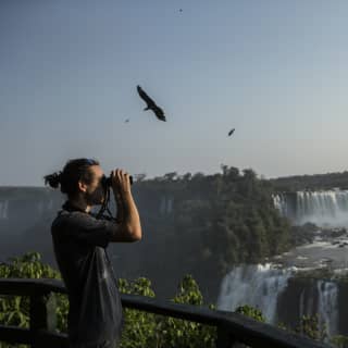 A guest looks through binoculars as large exotic birds circle above the Iguassu Falls at first light